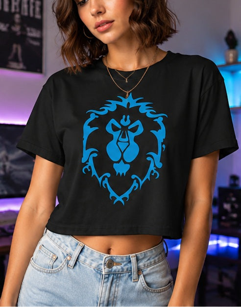 A woman wearing a black cropped t-shirt with an World of Warcraft Alliance Crest graphic on the front, paired with light wash blue jeans, set against a dimly lit gaming room background.